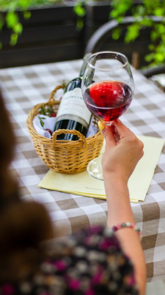 A person holds a glass of red wine at an outdoor table setting with a basket of wine bottles.